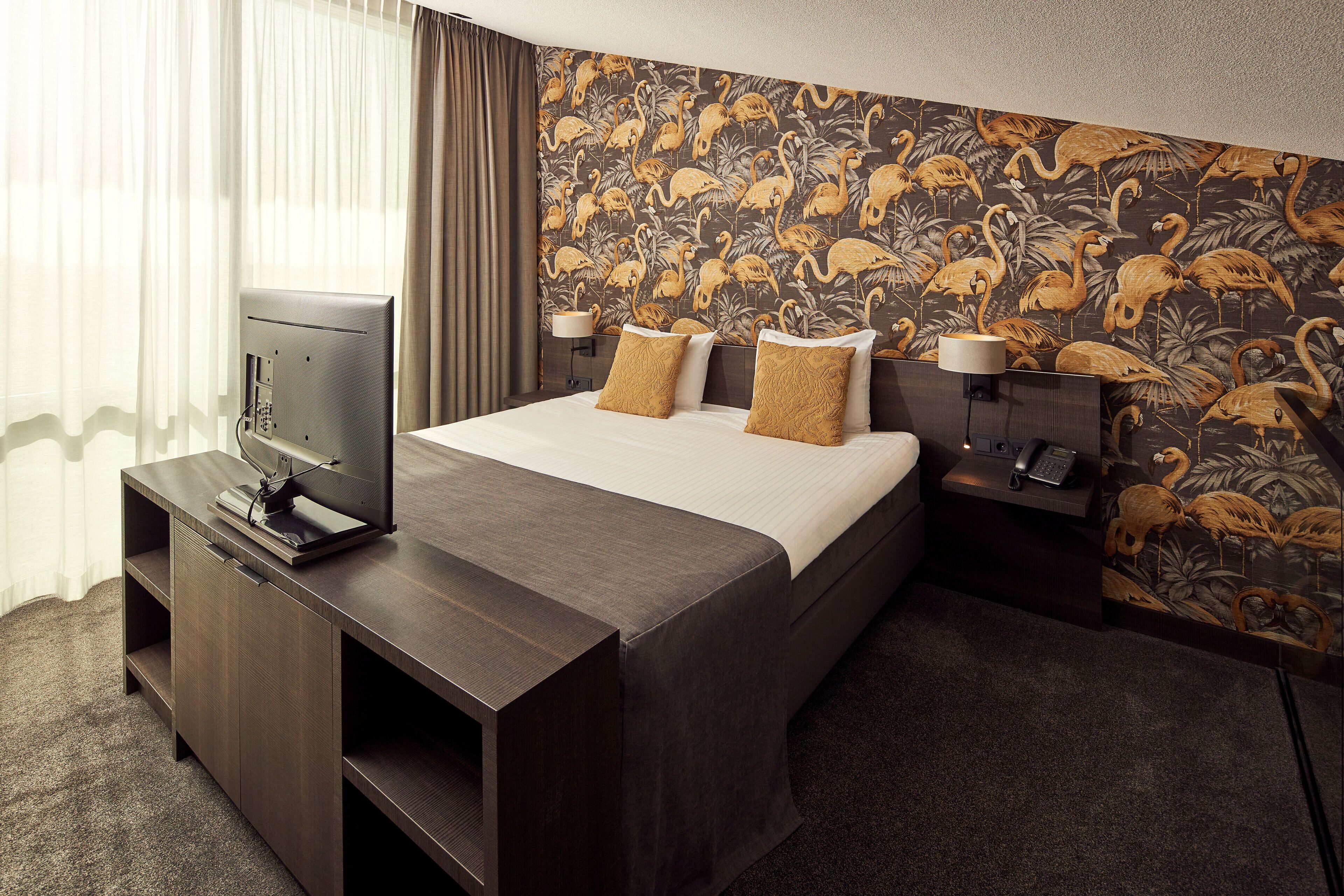 A hotel room with a bed, a TV on a stand, a desk with a lamp, and a wall with a tropical wallpaper.