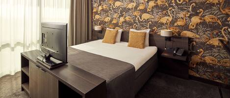 A hotel room with a bed, a TV on a stand, a desk with a lamp, and a wall with a tropical wallpaper.
