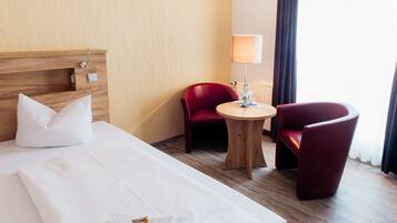 A hotel room with a bed, two red chairs, a small wooden table, a lamp, and a wall-mounted light switch.