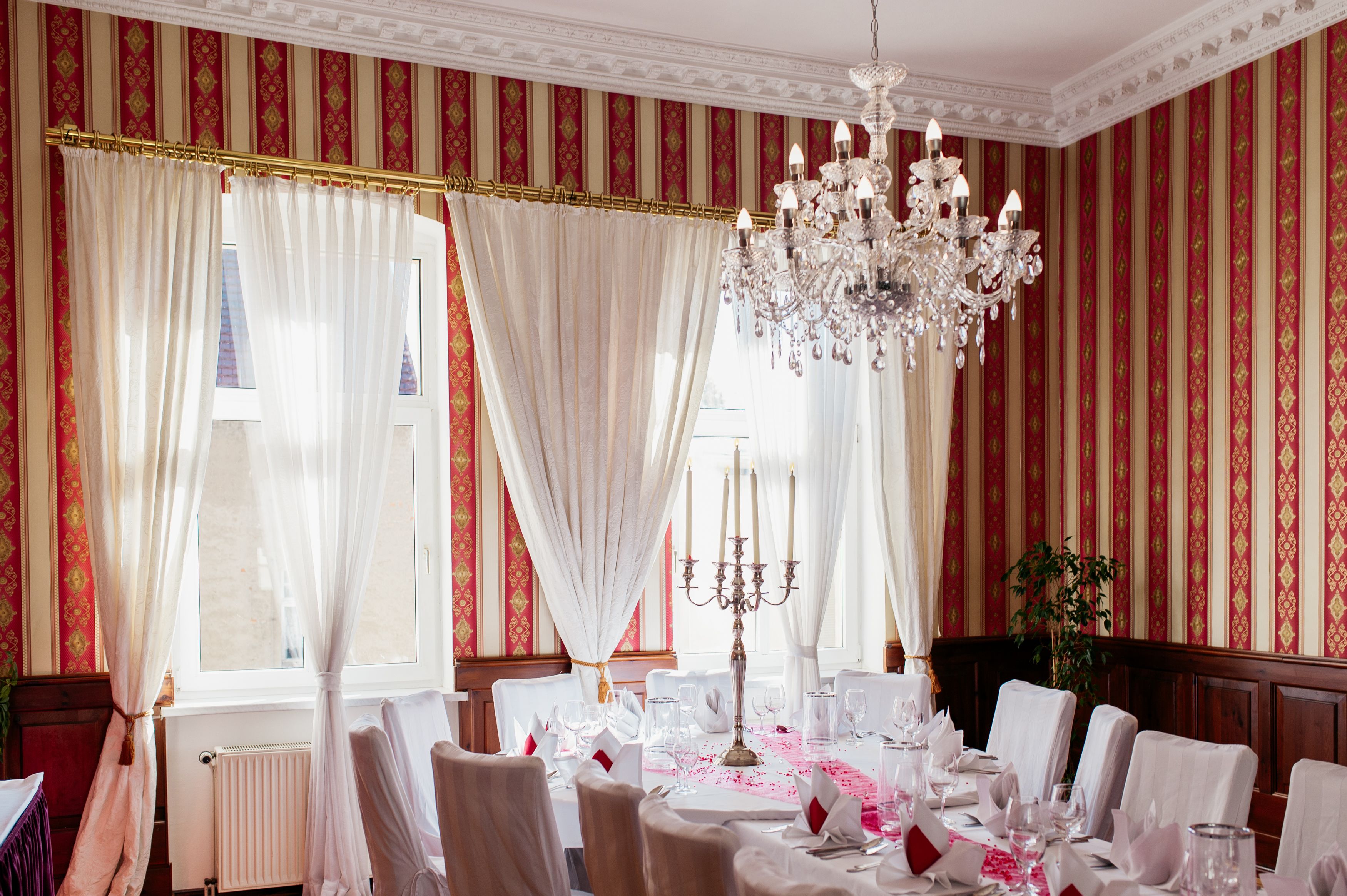 A formal dining room with a chandelier, white tablecloths, and red-striped wallpaper.