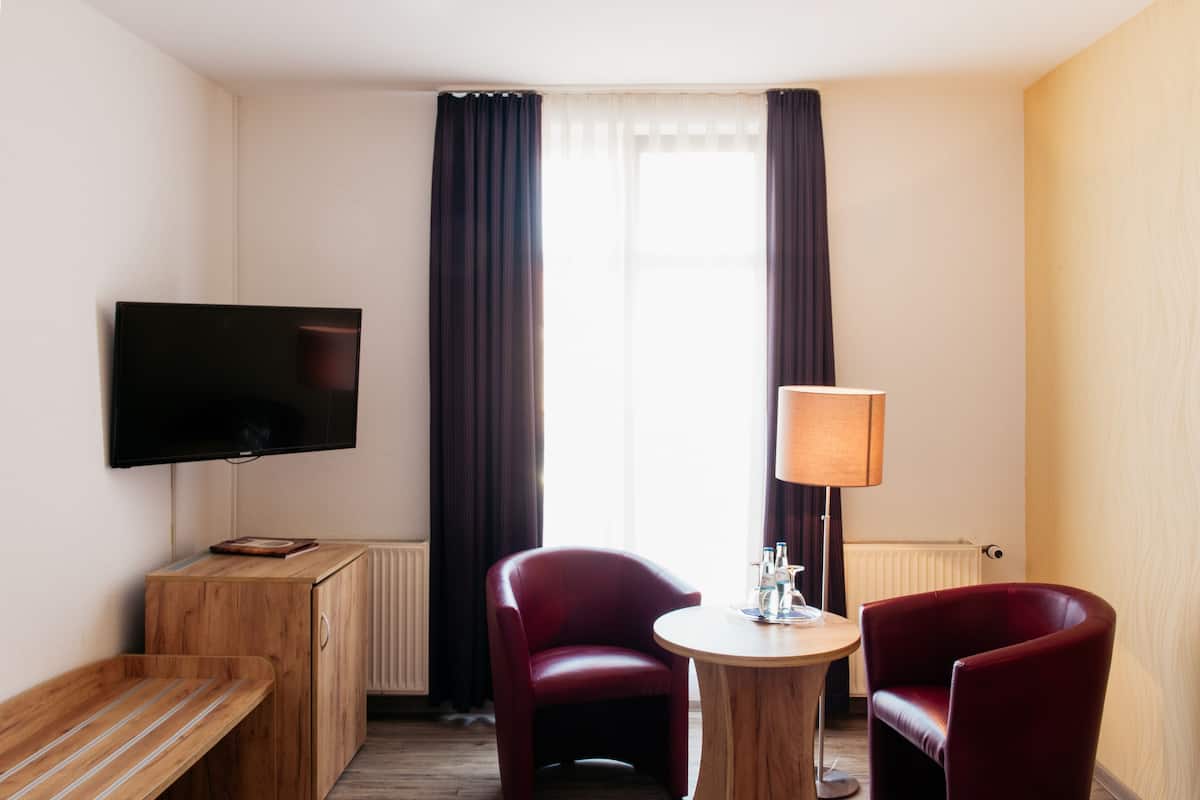 A hotel room with a flat-screen TV mounted on the wall, a wooden bench, two red chairs, a small round table with a lamp, and a window with dark curtains.