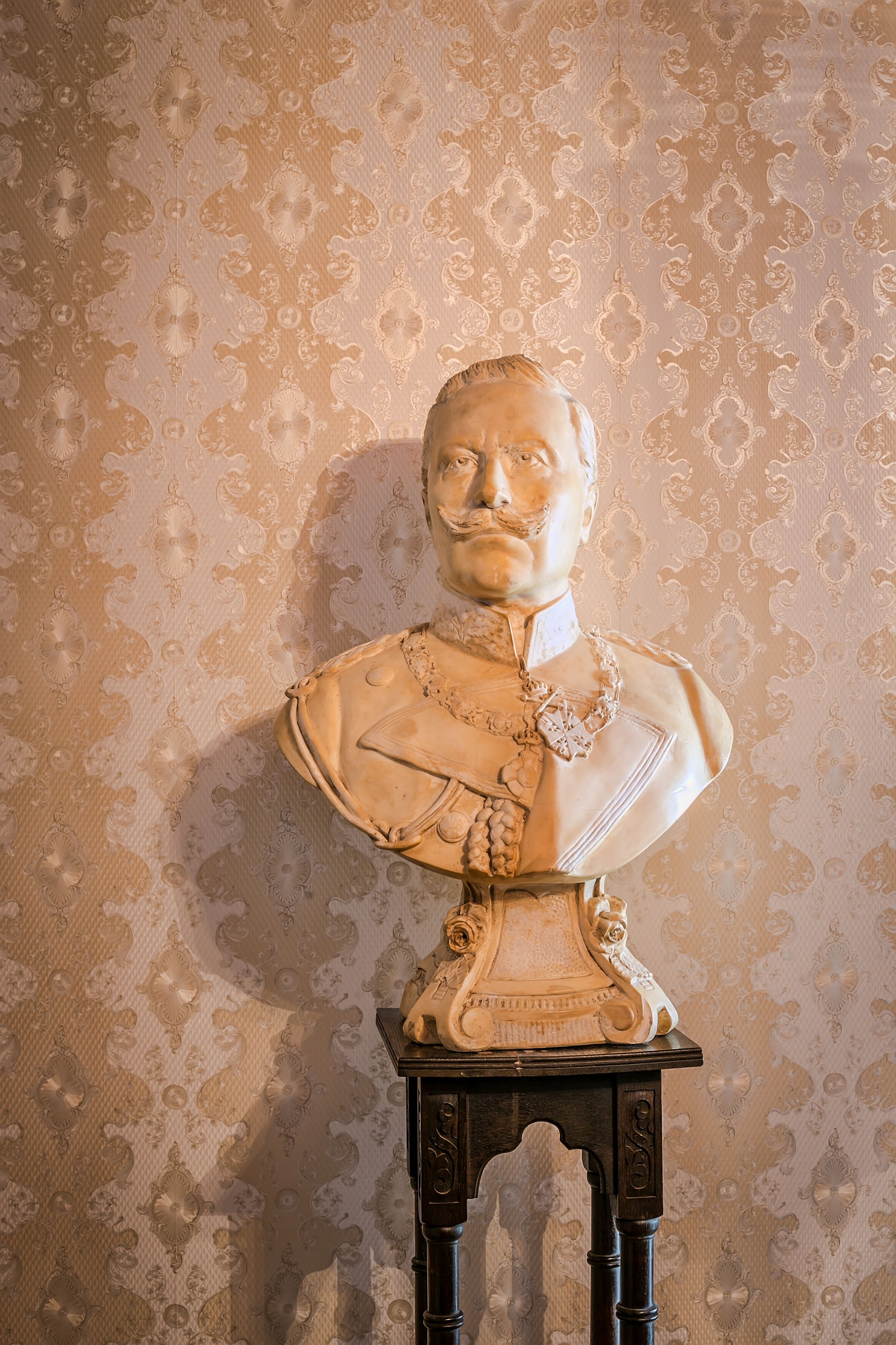 Una escultura de busto sobre un pedestal, frente a un fondo de papel tapiz con diseño.