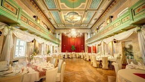 A banquet hall with ornate ceiling and chandeliers, multiple round tables set for dining, and a red curtain backdrop. - Hotel und Restaurant Deutscher Kaiser (Perleberg)