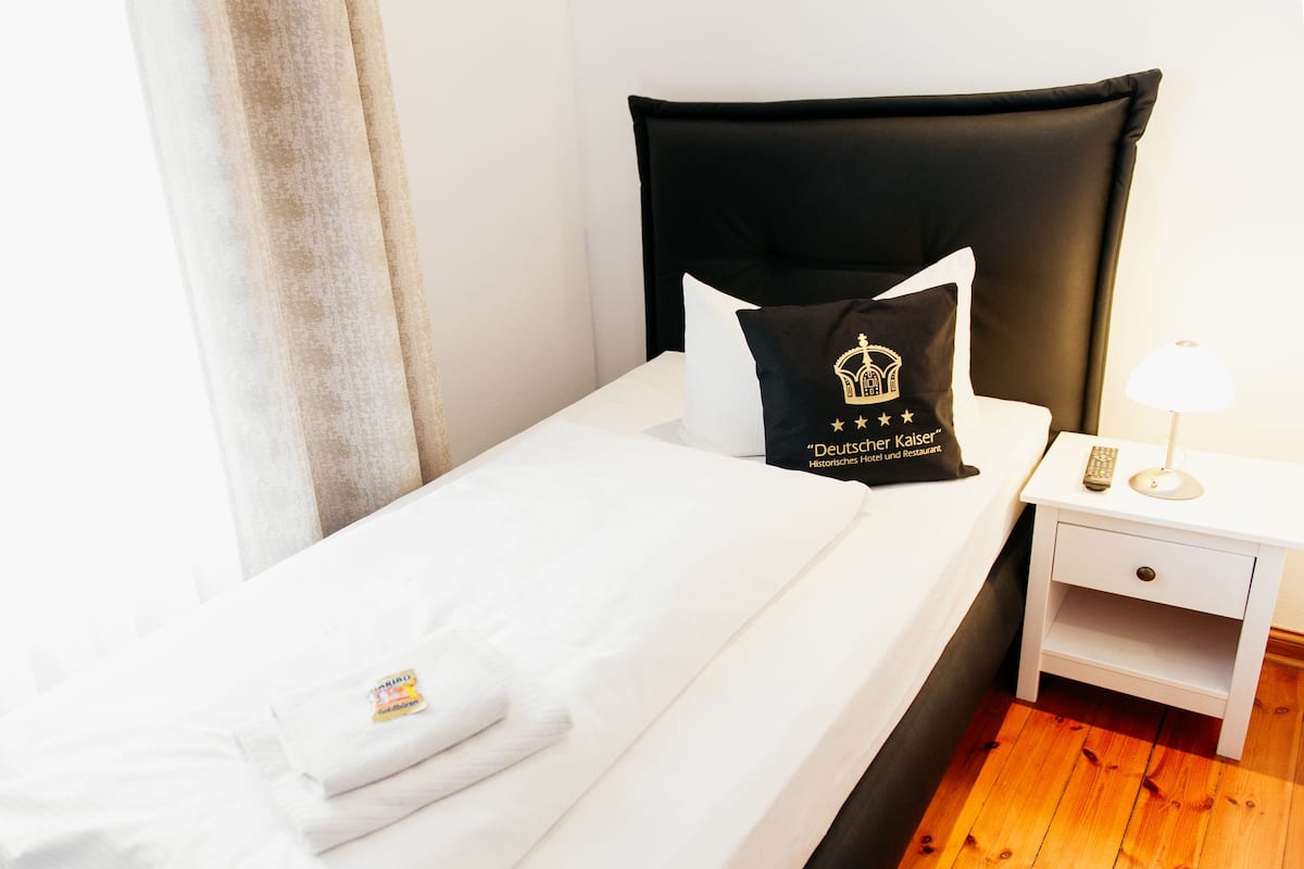 A neatly made bed with white linens and a black pillow with a logo. There is a white bedside table with a lamp and a remote control.