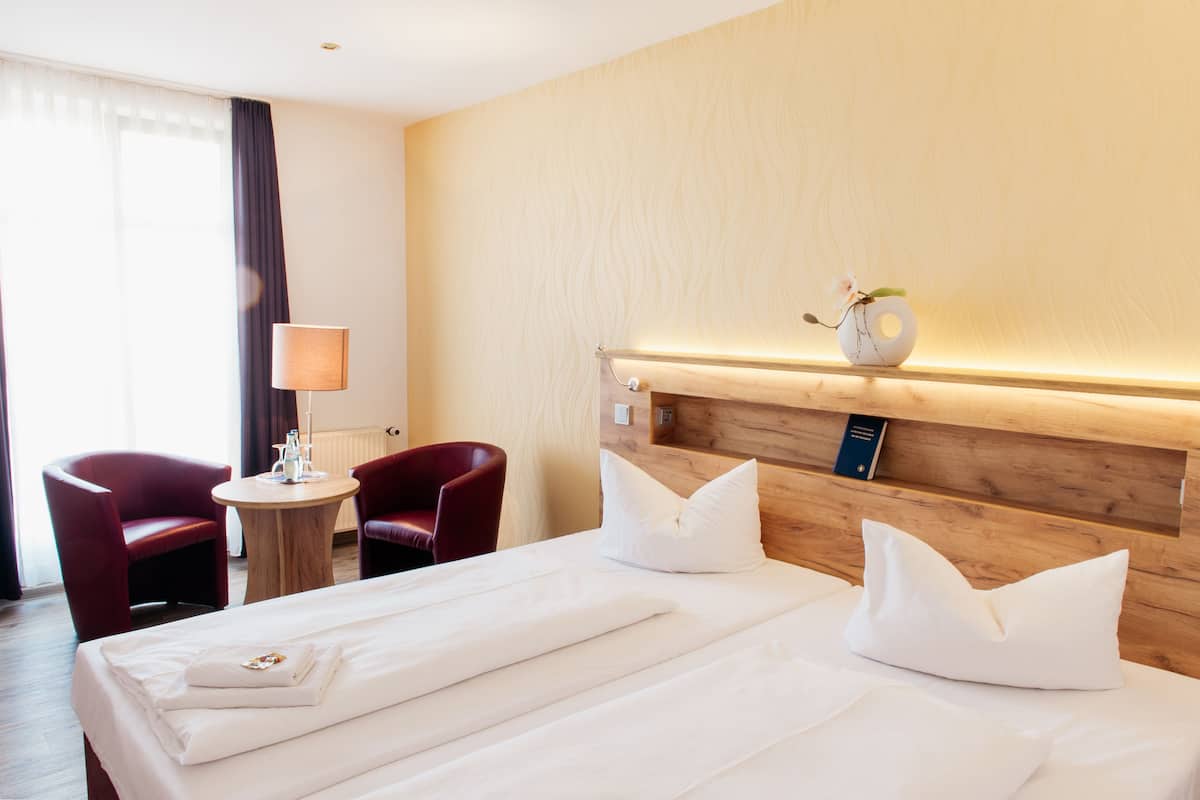 A hotel room with a wooden headboard, two single beds, a round side table with a lamp, and two red armchairs.