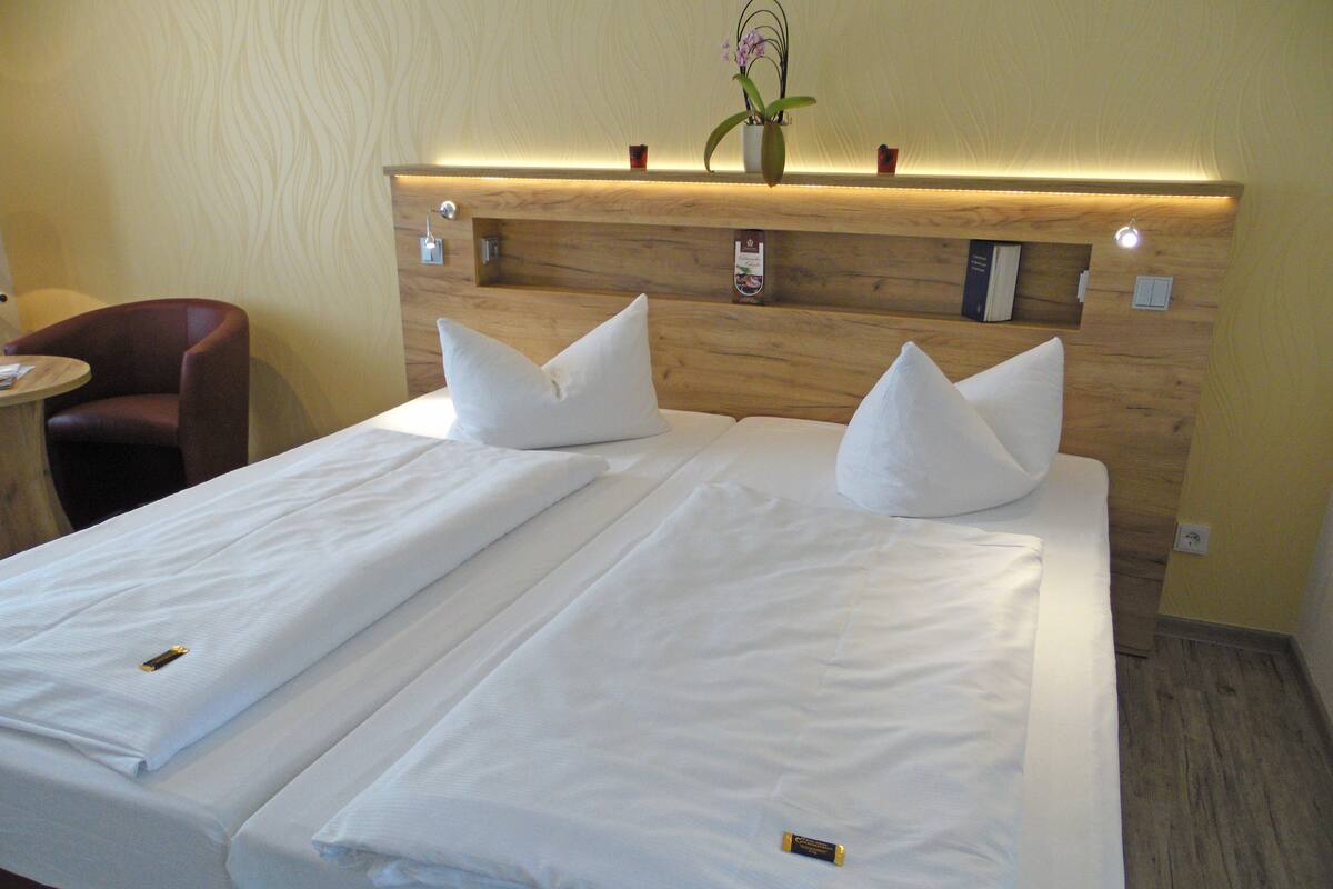 A hotel room with a double bed, two pillows, a bedside table, a chair, and a small table with a lamp.