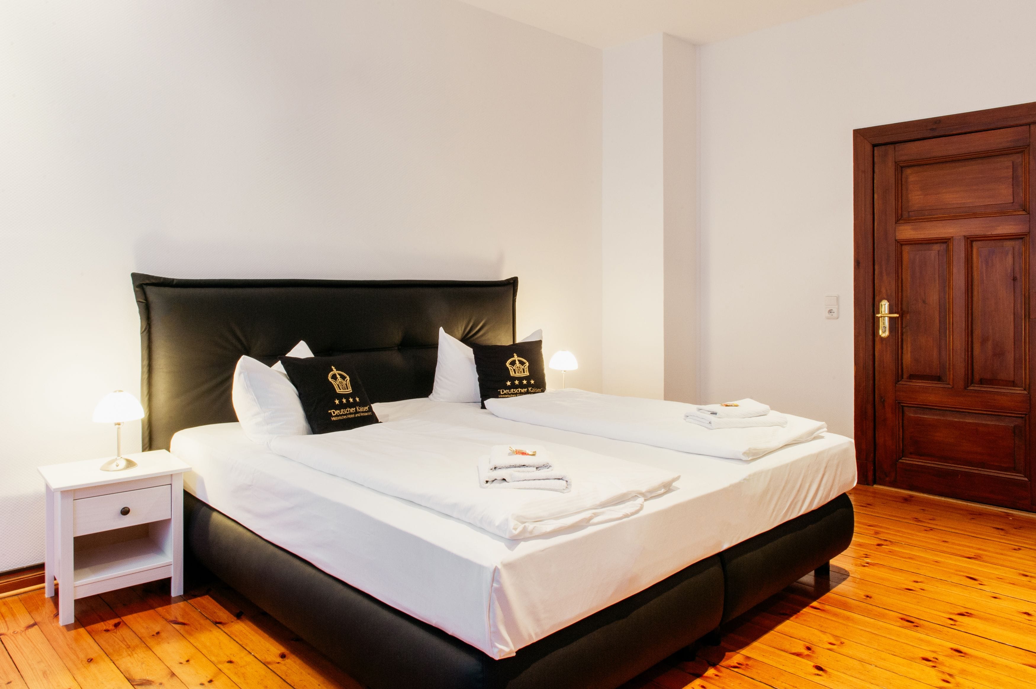 A double bed with black headboard and white bedding, a nightstand with a lamp, and a wooden door.