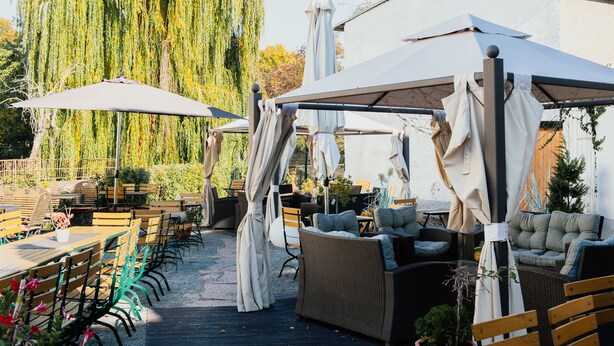 An outdoor patio area with wooden tables and chairs, a covered seating area, and a weeping willow tree. - Hotel und Restaurant Deutscher Kaiser (Perleberg)