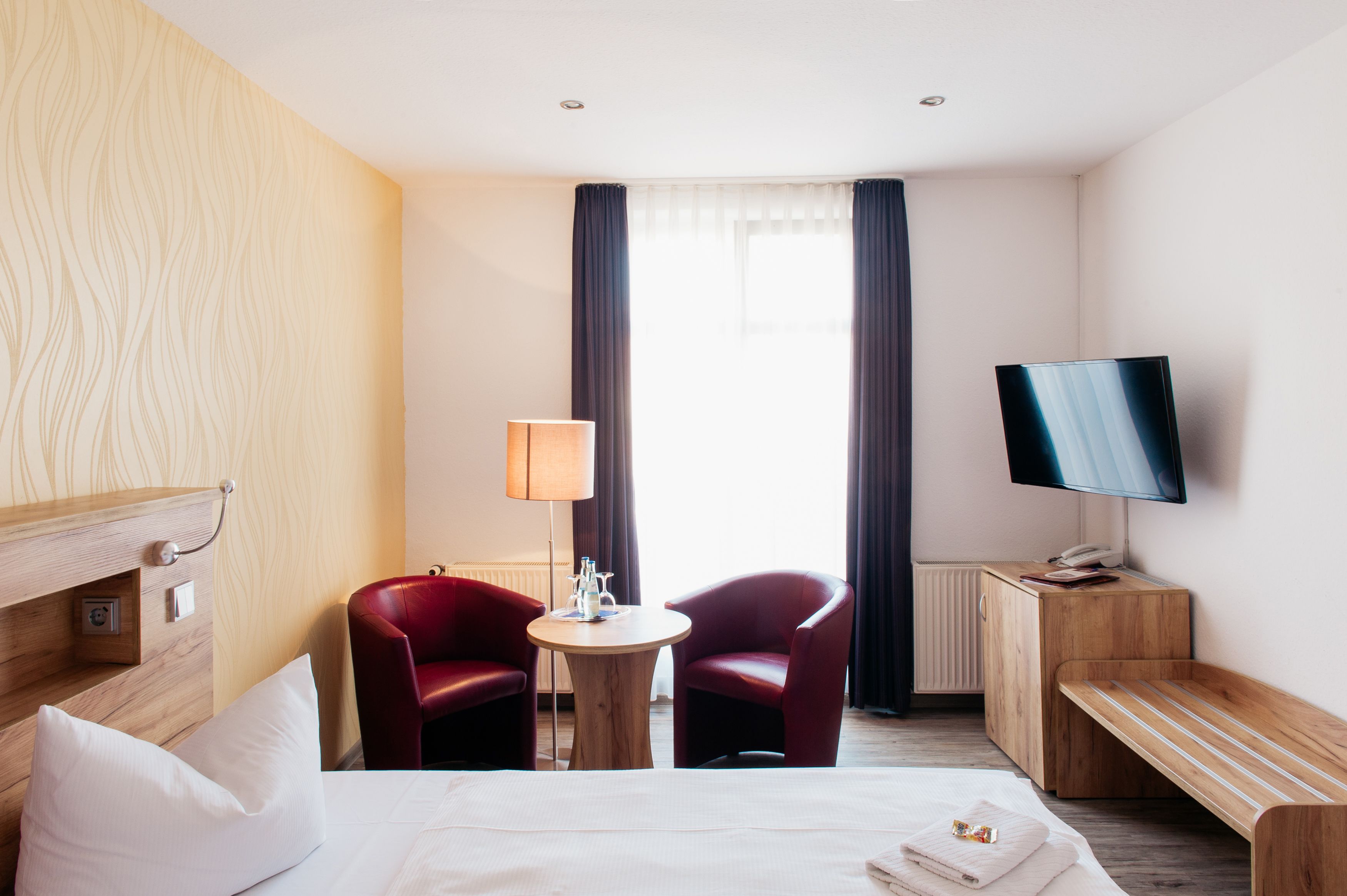 A hotel room with a bed, two red chairs, a small round table, a lamp, a TV, and a wooden bench.