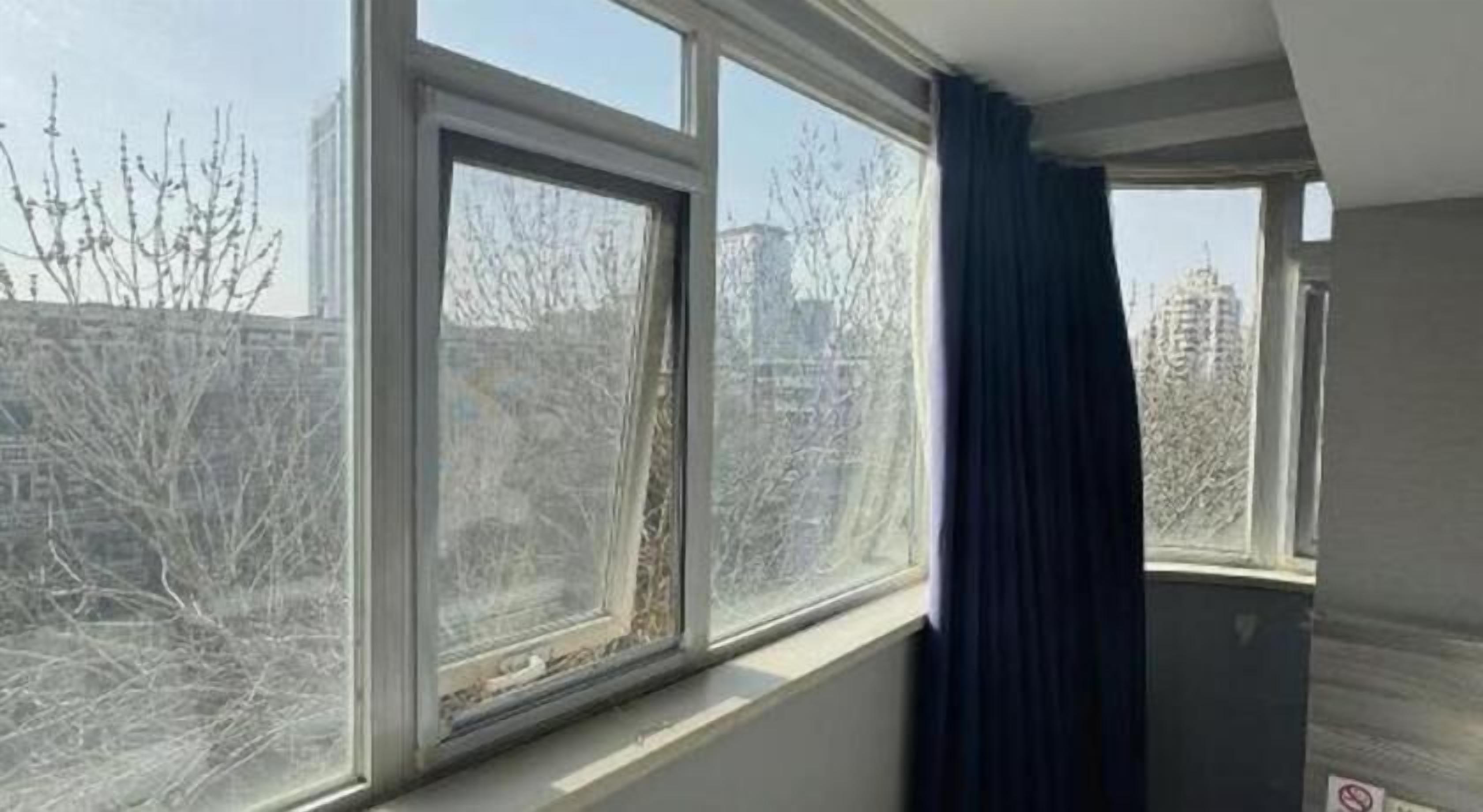 A room with a large window, a dark curtain, and a view of trees and buildings outside.