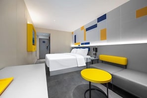 A modern hotel room with a bed, a seating area with a yellow bench, a small round table, and a wall with geometric patterns.