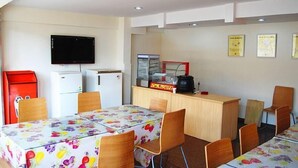 A dining area with tables covered in floral tablecloths, wooden chairs, a television mounted on the wall, a refrigerator, and a coffee machine. - 7 Days Inn (Tianjin)