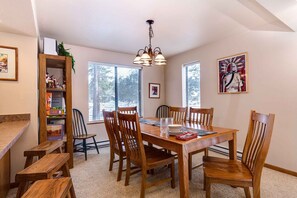 Un coin repas avec une table en bois, des chaises et un lustre. On y trouve des fenêtres avec des stores, une bibliothèque et des œuvres d’art encadrées accrochées aux murs.