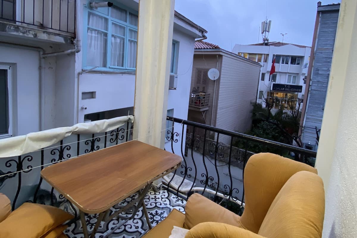 A balcony with a wooden table and yellow armchairs, overlooking a residential area with white buildings and a red flag.