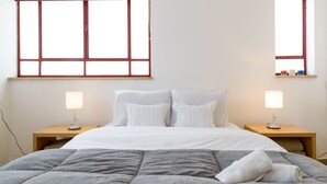 Una cama bien hecha con un edredón gris, dos lámparas de noche y una ventana con marco de madera.
