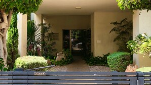 Una entrada de edificio con un camino de acceso porticado, un pequeño jardín con diversas plantas y un balcón con macetas.