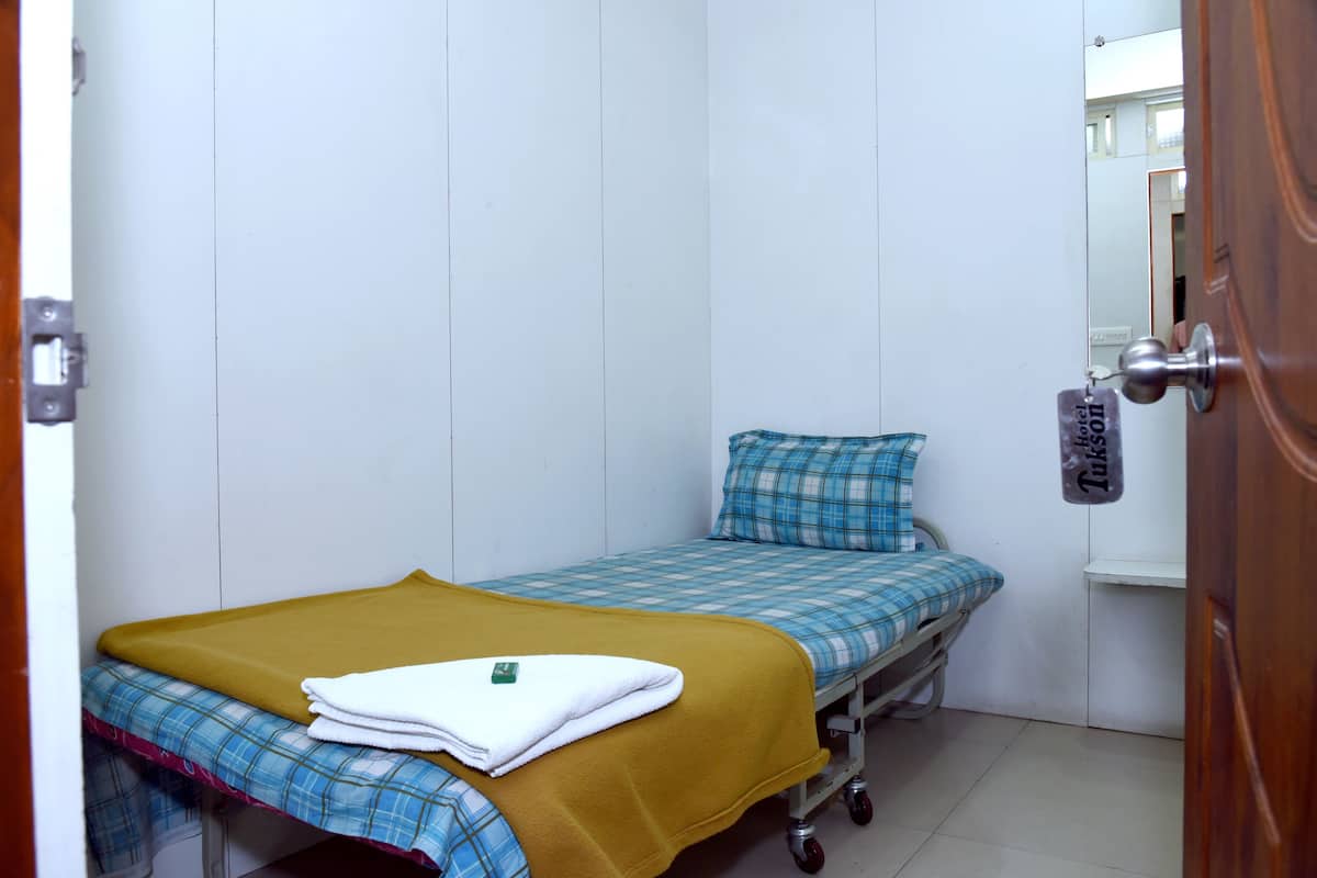 Una habitación pequeña con una cama individual, una puerta de madera, una colcha azul y blanca a cuadros, una manta color mostaza y una toalla blanca.