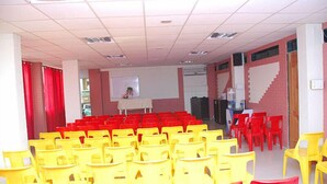 Un salón de conferencias con filas de sillas amarillas y rojas, una pizarra y una pantalla para proyector.