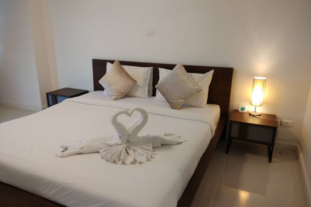 A neatly made bed with a heart-shaped towel arrangement, white bedding, and a bedside lamp on a wooden nightstand.