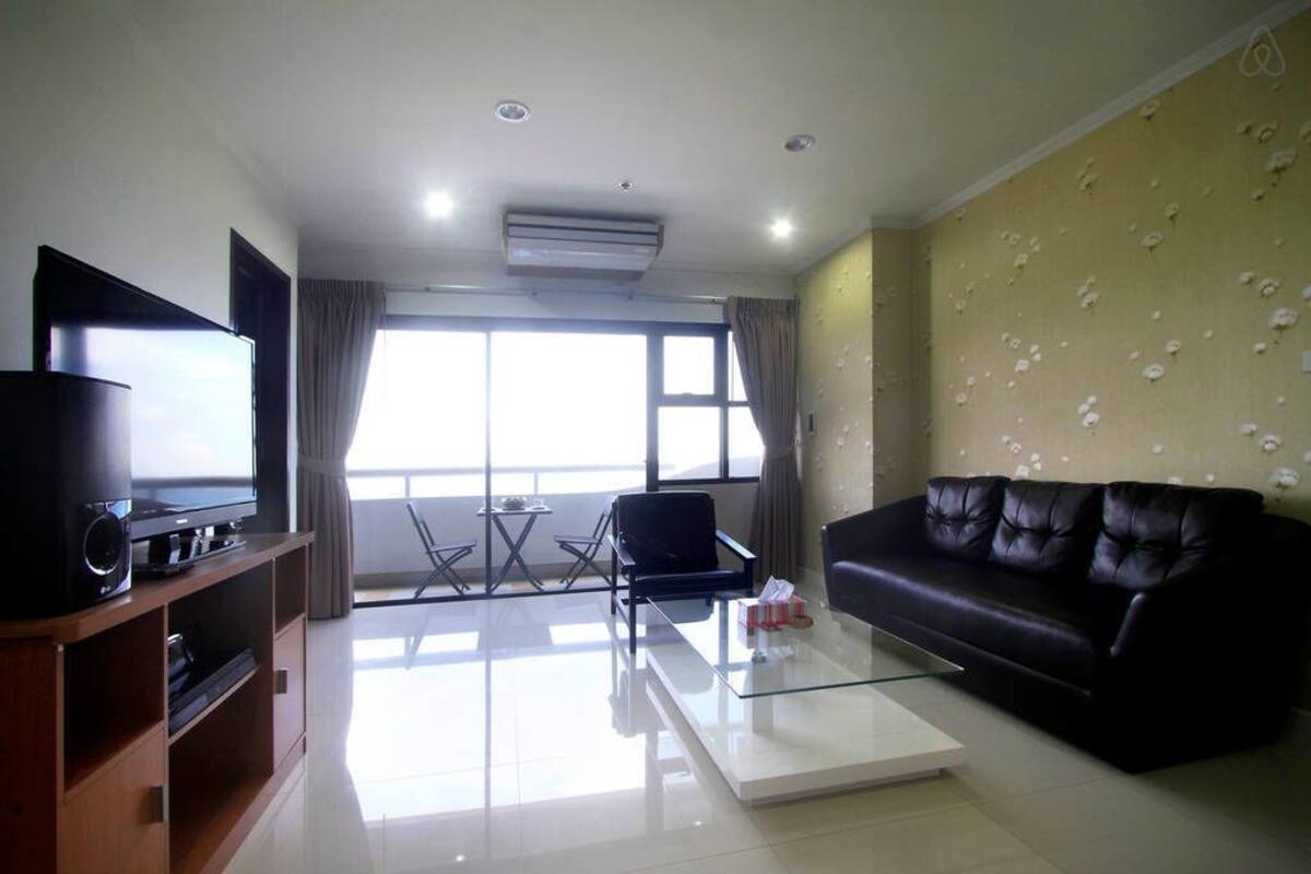 A modern living room with a flat-screen TV, leather sofa, glass coffee table, and a large window with curtains.