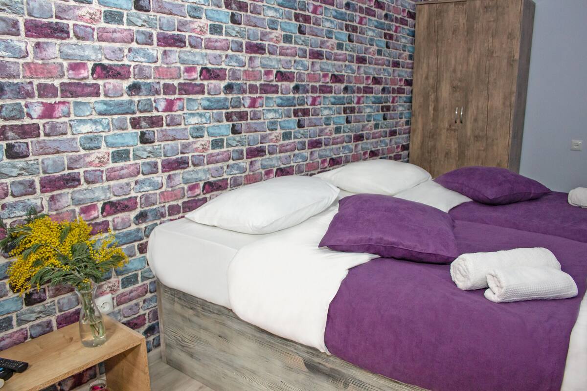 A bedroom with a brick-patterned wall, a wooden wardrobe, a bed with white and purple bedding, and a small table with a vase of flowers.
