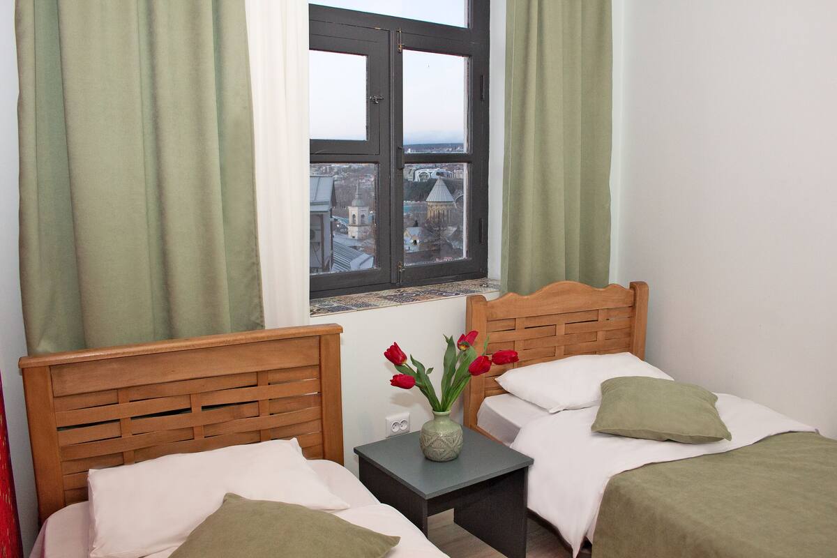 A room with two beds, a window with a view of a cityscape, and a vase with flowers on a nightstand.
