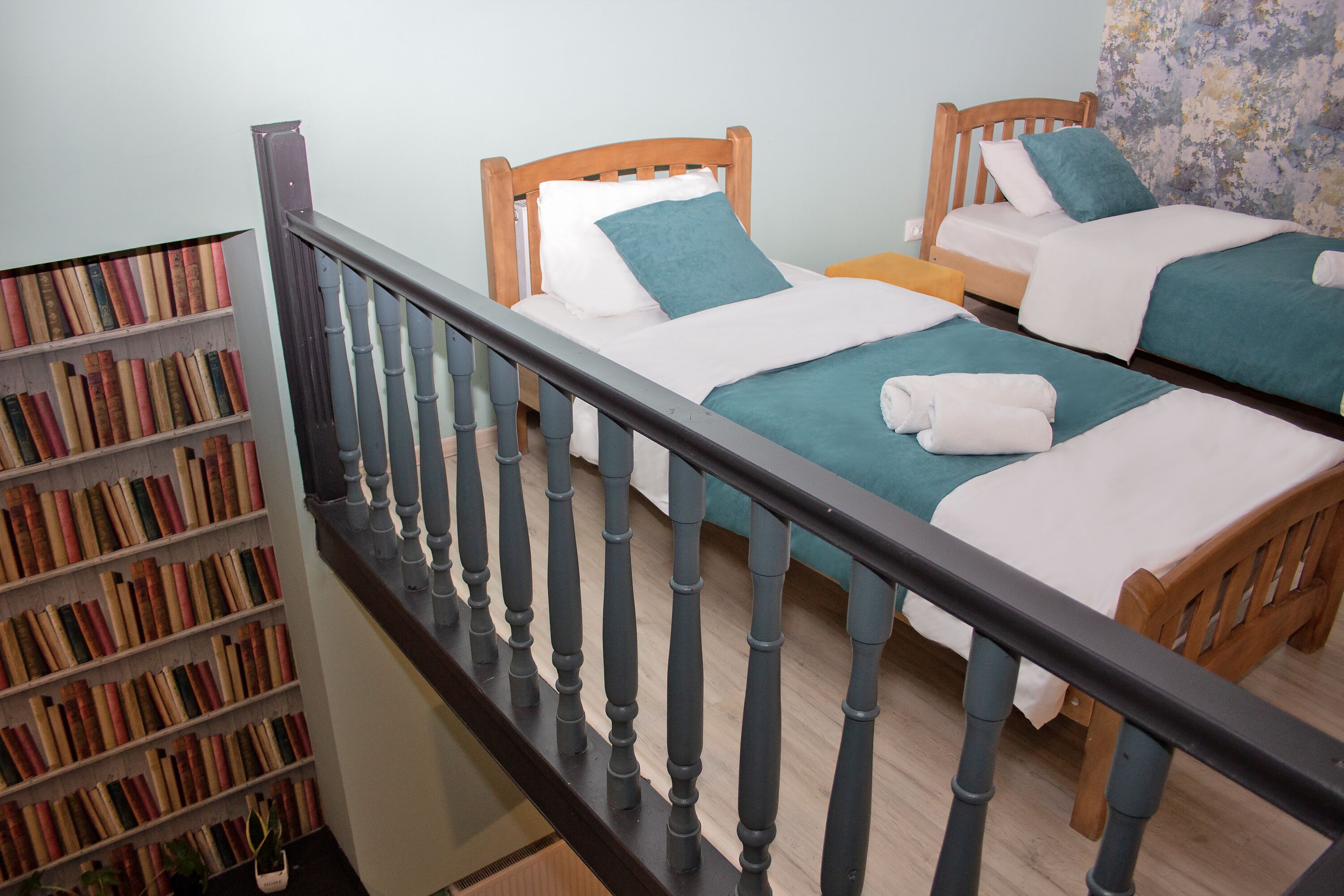 A staircase leading to a room with two wooden beds, each with a blue bedspread and white pillows. A bookshelf filled with books is visible to the left.