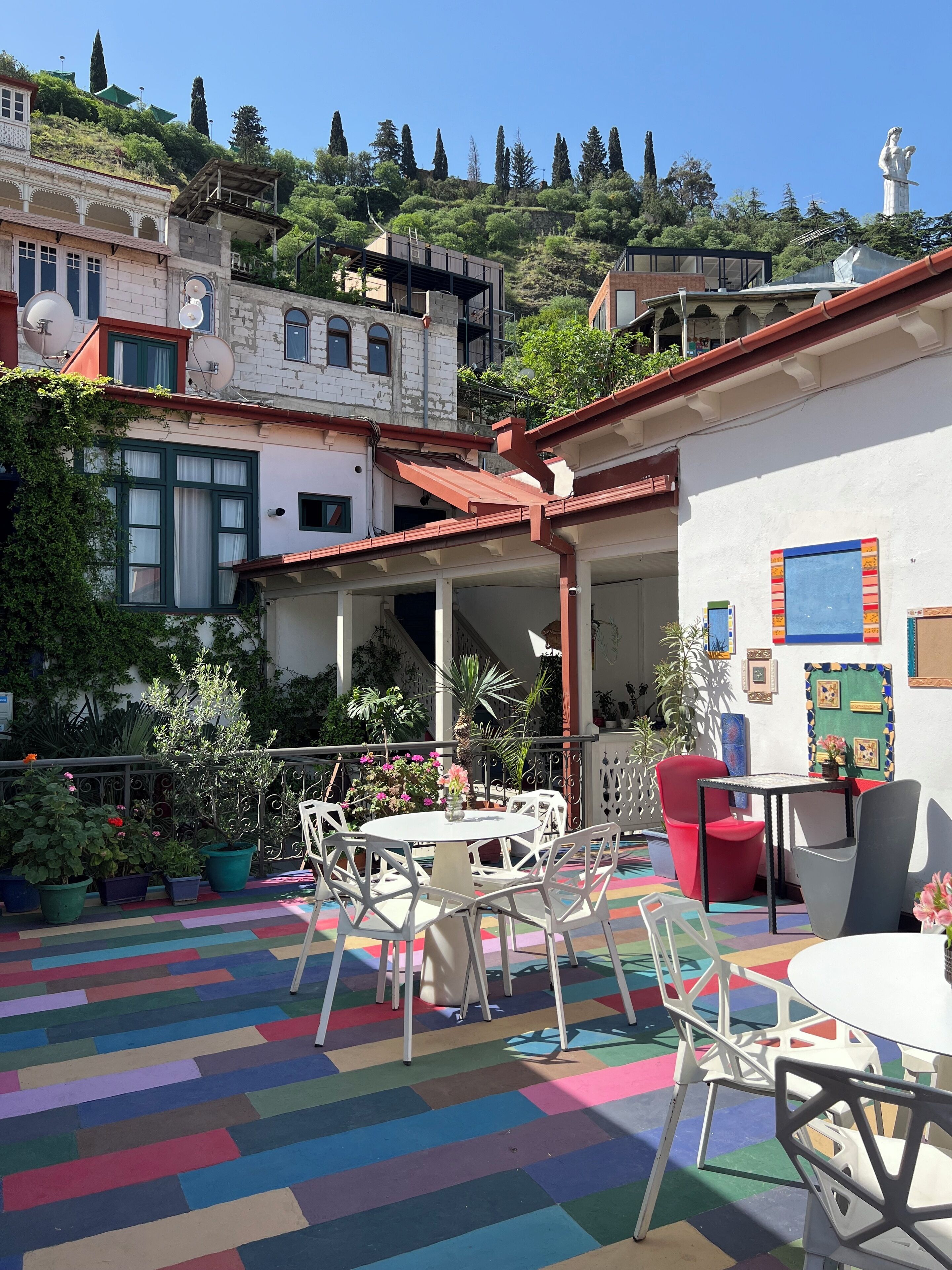 Un patio con piso colorido, sillas y mesas blancas, rodeado de edificios y vegetación.