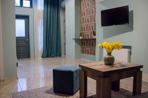 Moderni olohuone, jossa on puinen pöytä, tummansininen jalkarah, kirjahylly ja seinälle asennettu taulutelevisio.