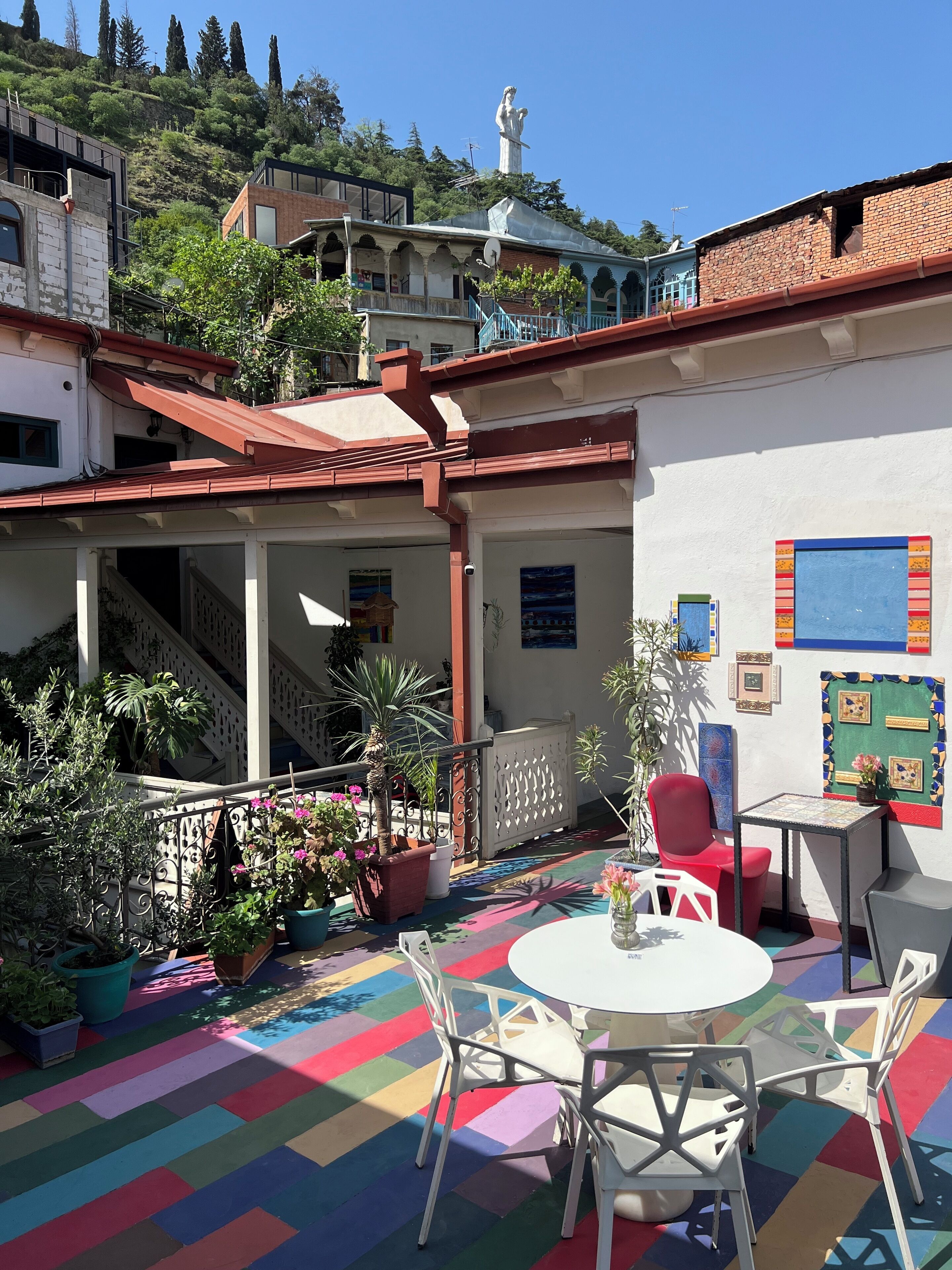 A courtyard with a colorful floor, white chairs, a small table, and potted plants.