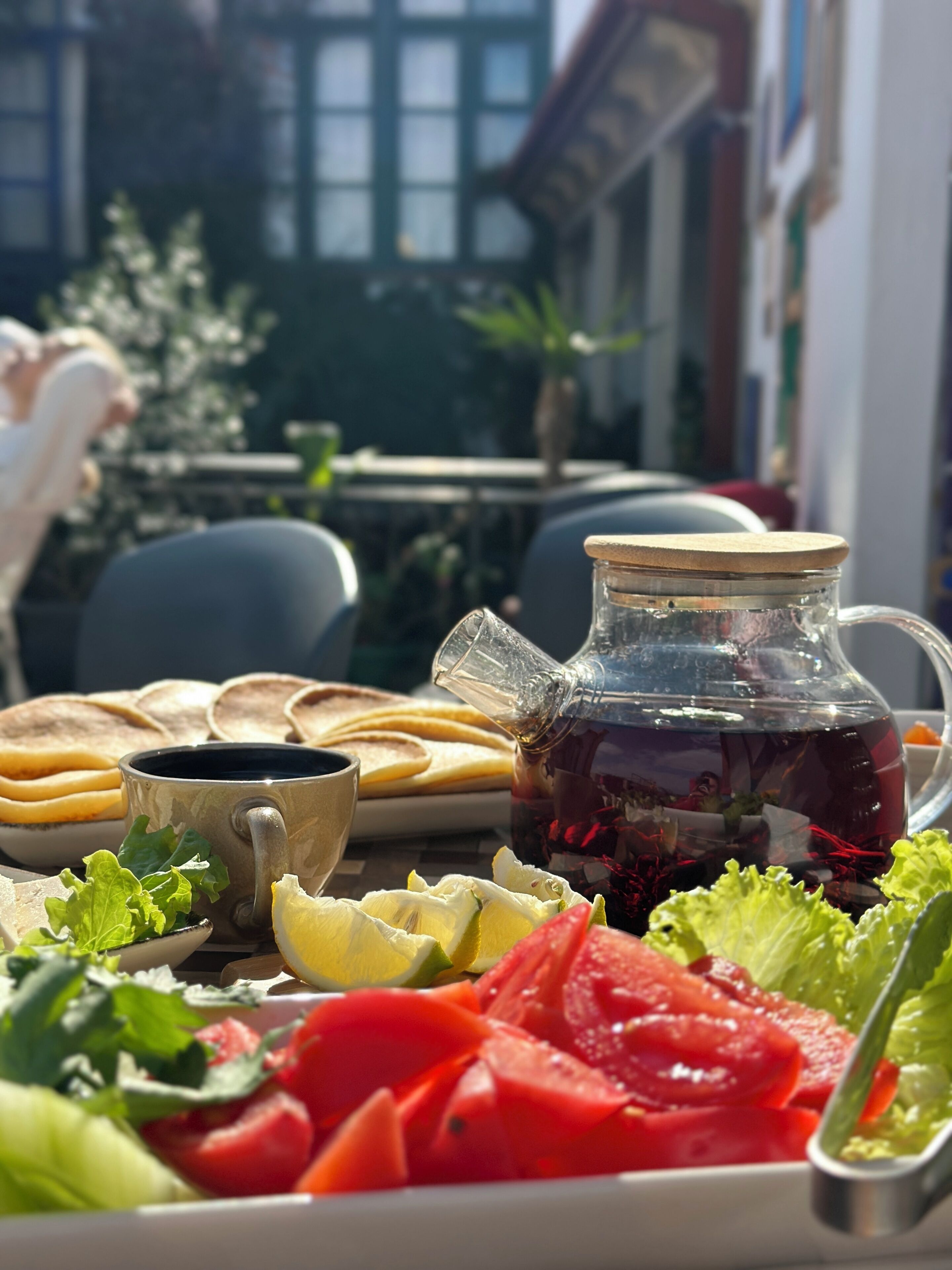 A table with a pitcher of dark liquid, possibly tea, and a cup, surrounded by various food items like sliced tomatoes and greens.