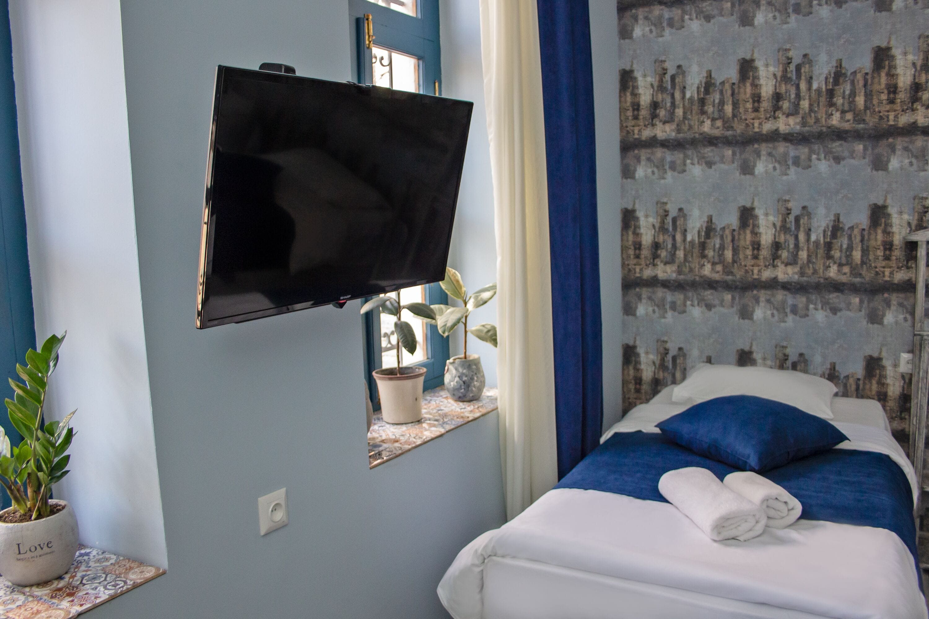 Une chambre d’hôtel avec un lit, un téléviseur fixé au mur, une fenêtre avec des rideaux et une plante en pot.