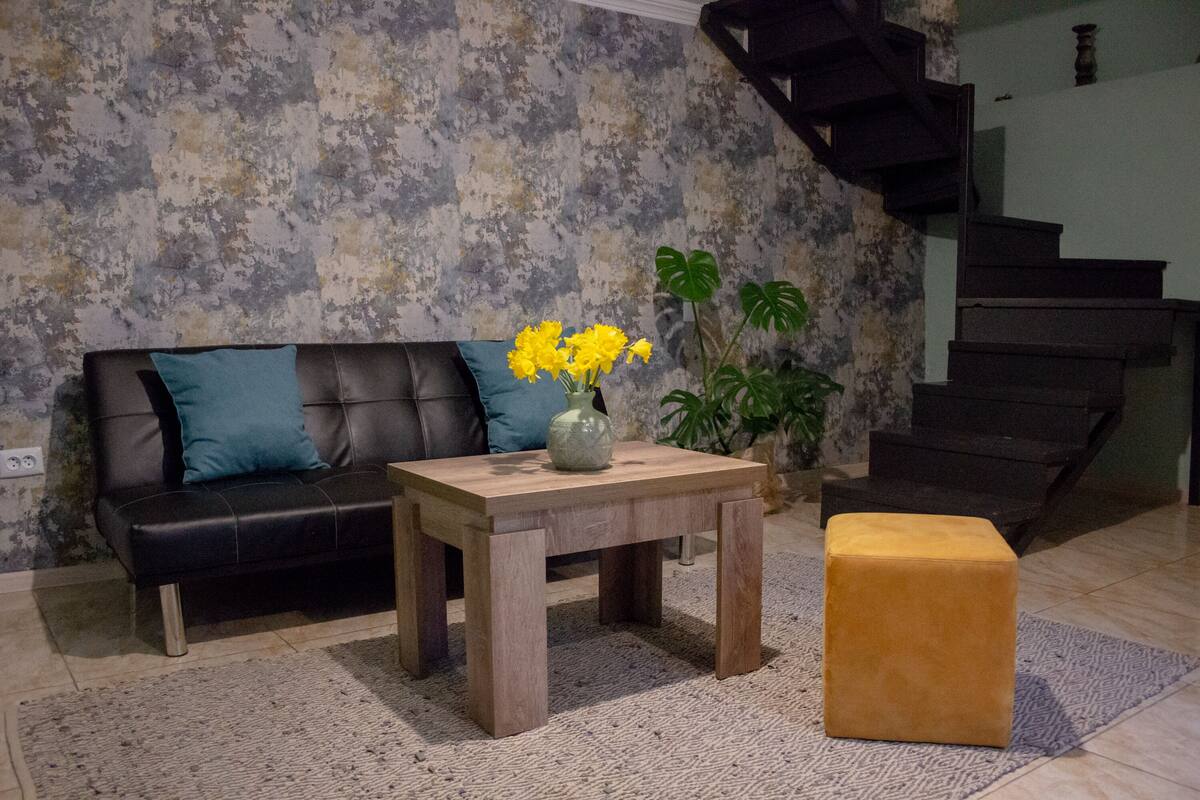 A living room with a black leather sofa, a wooden coffee table with a vase of yellow flowers, a potted plant, and a staircase in the background.