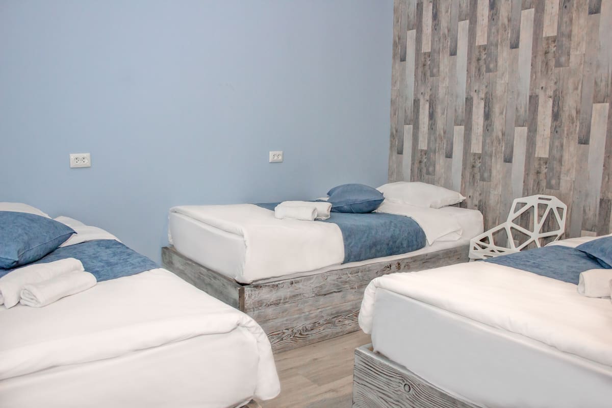 A hotel room with three beds, each with white bedding and blue accents. The room has a light blue wall and a wooden accent wall.