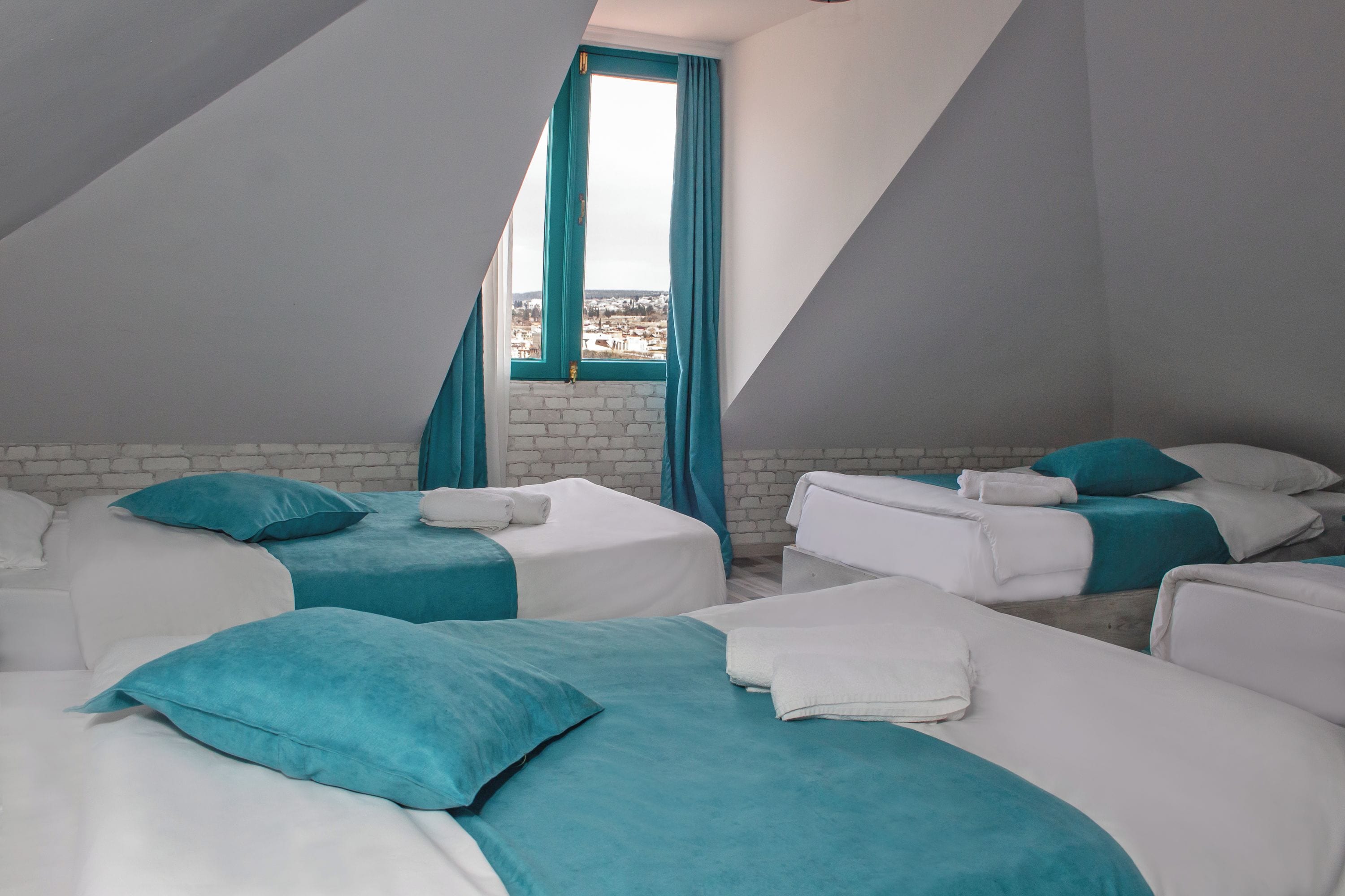 A room with two beds, each with a teal bedspread and white pillows, a white brick wall, and a window with teal curtains.