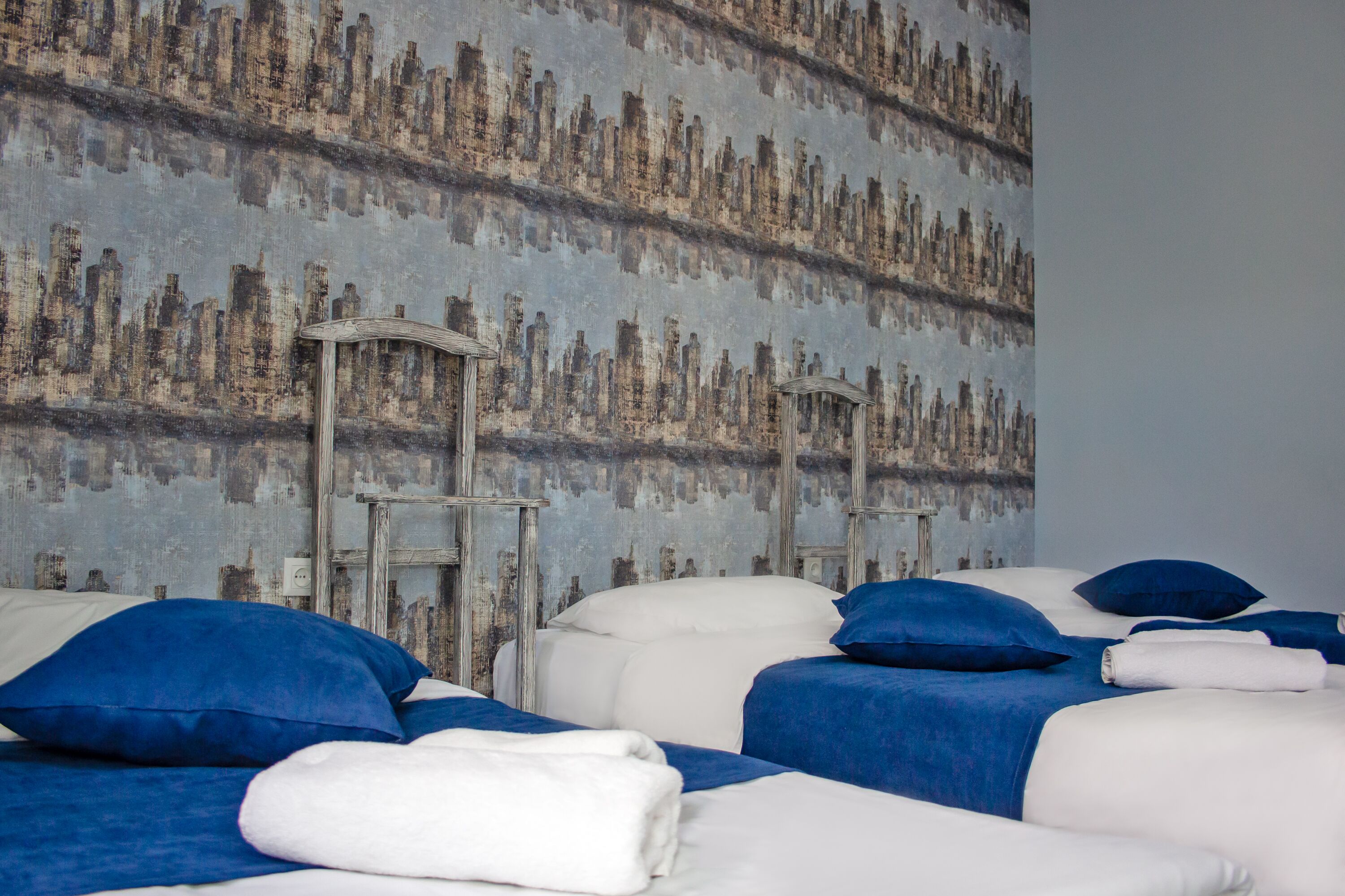 A hotel room with two beds, each with blue and white bedding, and a large wall mural of a cityscape.