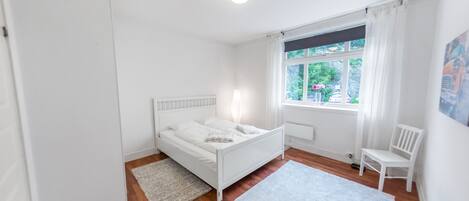 A white bedroom with a bed, a chair, a window with curtains, and a radiator.