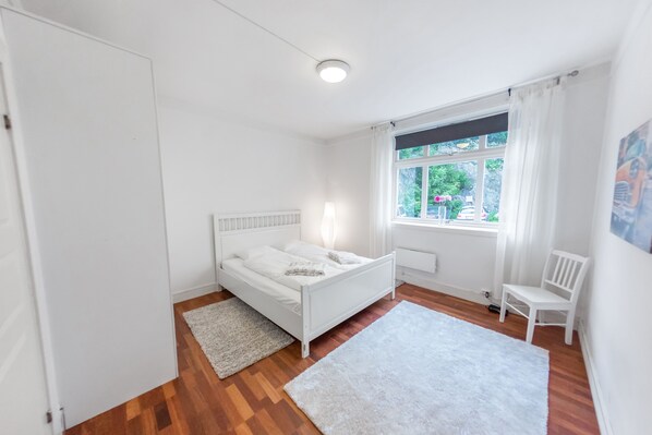 A white bedroom with a bed, a chair, a window with curtains, and a radiator.
