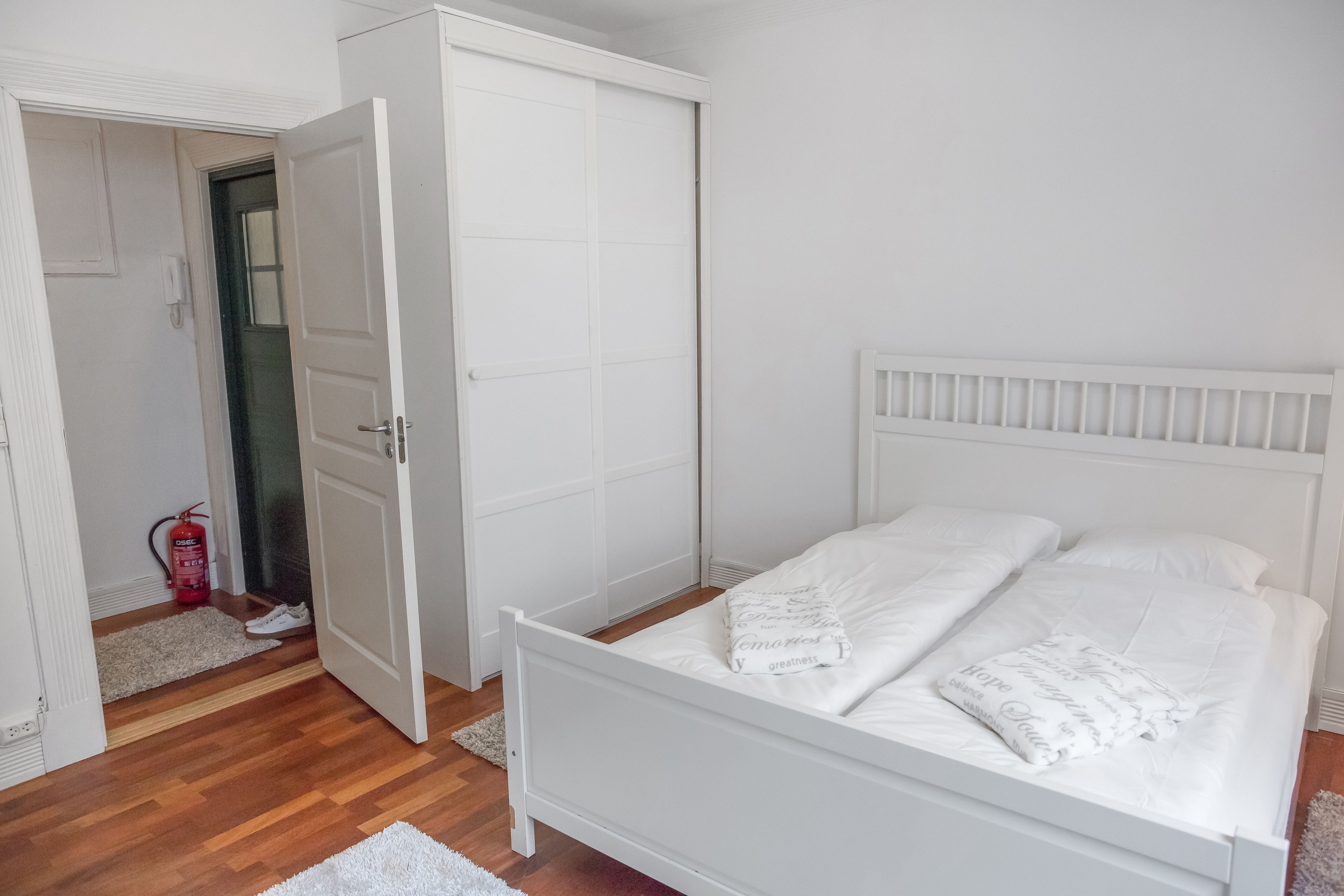 A bedroom with a white bed, a closed door, and a fire extinguisher in the corner.