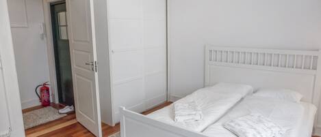 A bedroom with a white bed, a closed door, and a fire extinguisher in the corner.
