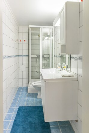 A bathroom with white tiles, a glass shower enclosure, a white sink, and a blue rug.