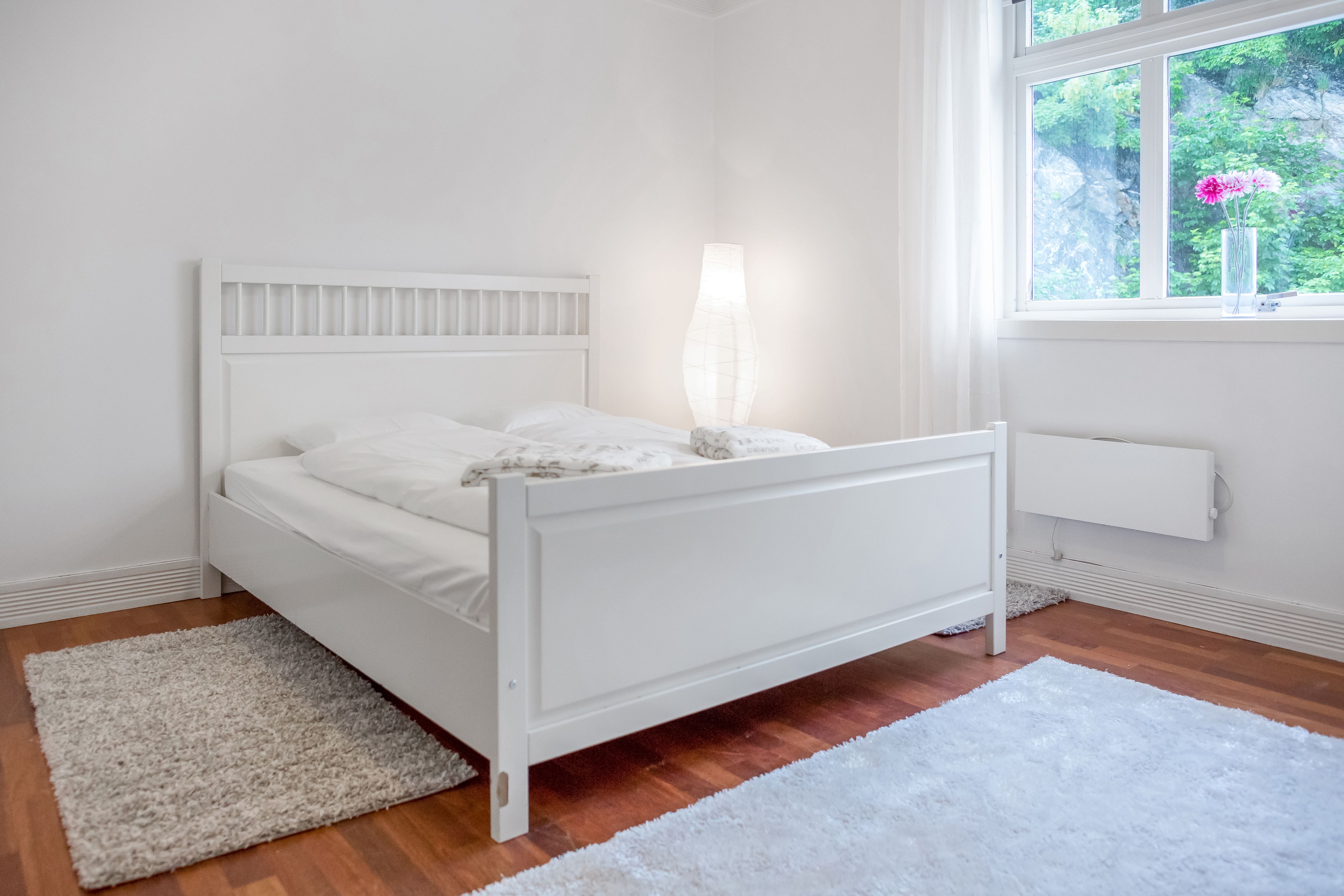 A white bed with a headboard, a bedside lamp, and a window with a view of greenery.