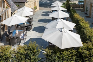 An outdoor patio area with multiple tables and white umbrellas, surrounded by stone walls and greenery. - The Lion Wendlebury (Bicester)