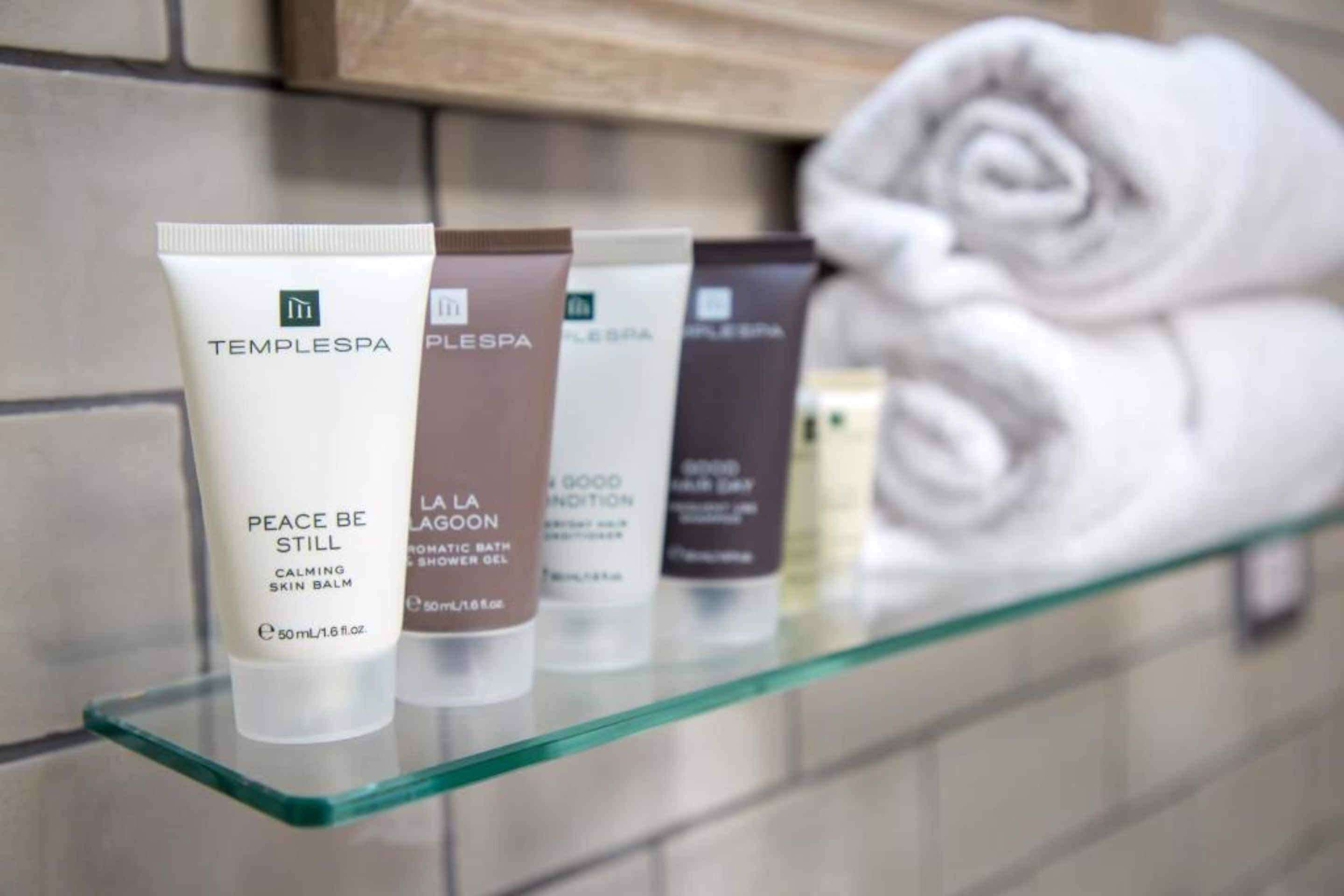 Uma prateleira de vidro com produtos de higiene pessoal, incluindo um bálsamo calmante para a pele, um gel de banho e um hidratante corporal em boas condições.