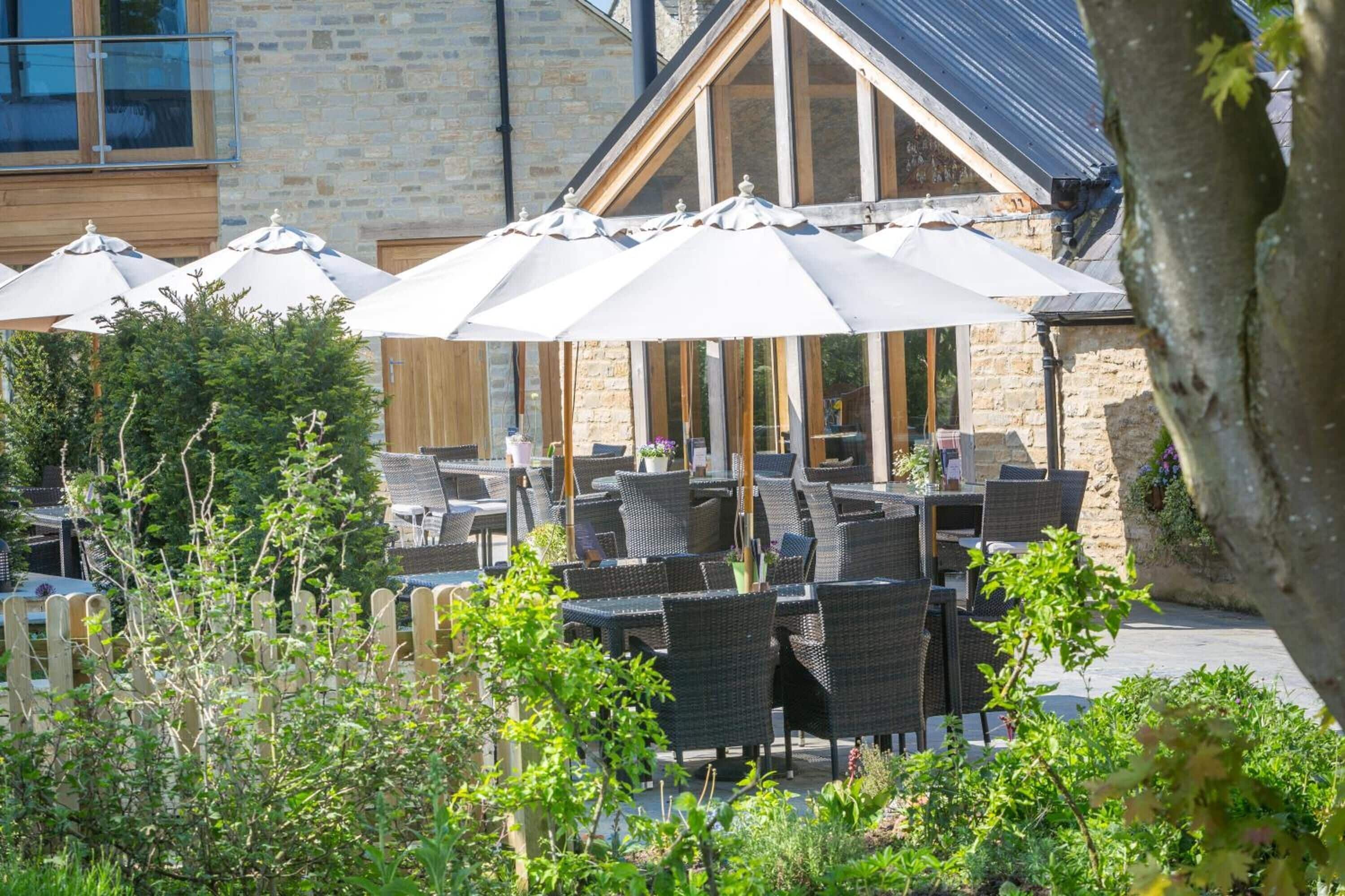 An outdoor seating area with wicker tables and chairs, large umbrellas, and a stone building in the background.