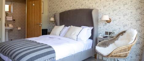 A bedroom with a bed, a chair, a nightstand, a lamp, and a bathroom visible through an open door.