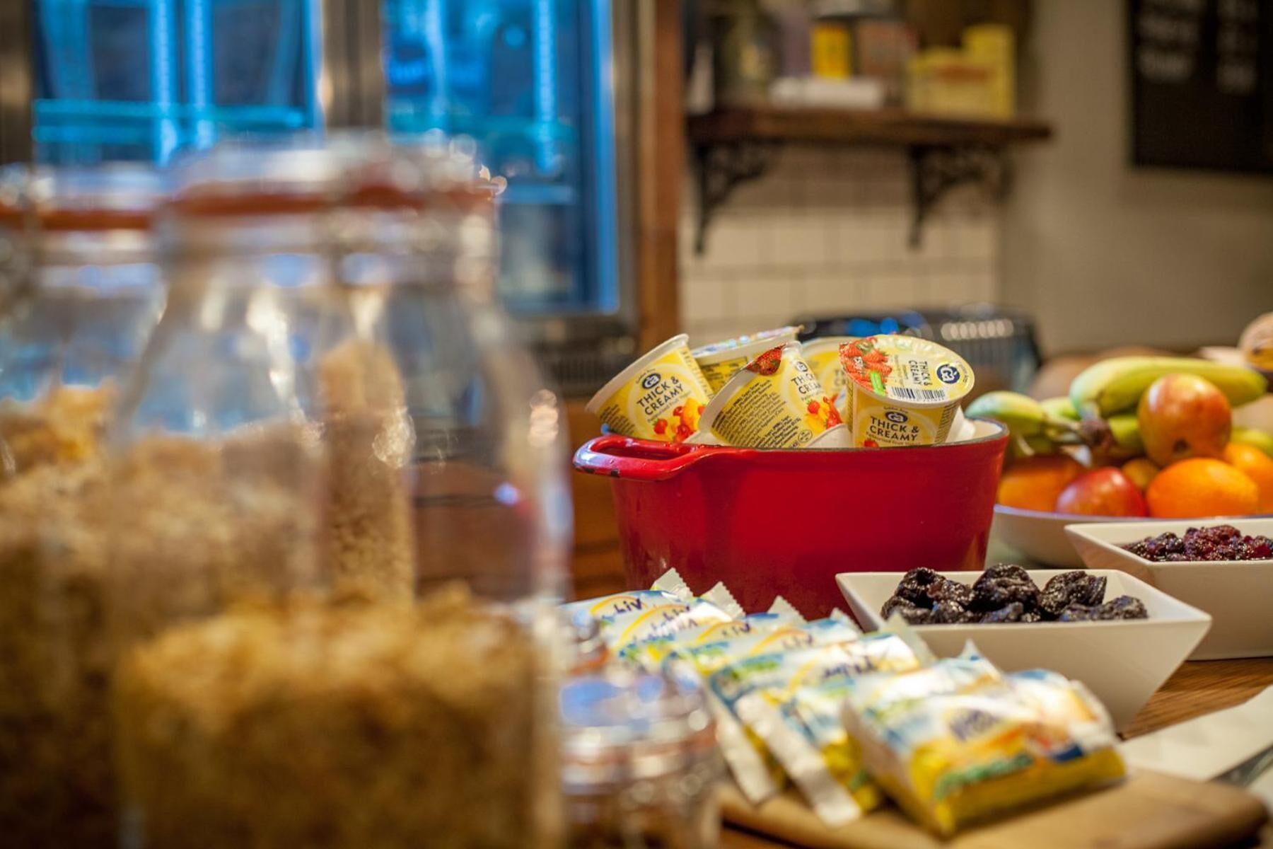 Una encimera de cocina con frascos, un tazón rojo lleno de vasos de yogur y diversas frutas y aperitivos envasados.