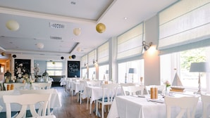 A restaurant with white tables and chairs, large windows, and decorative hanging lights. - Waastwinj (Utersum)