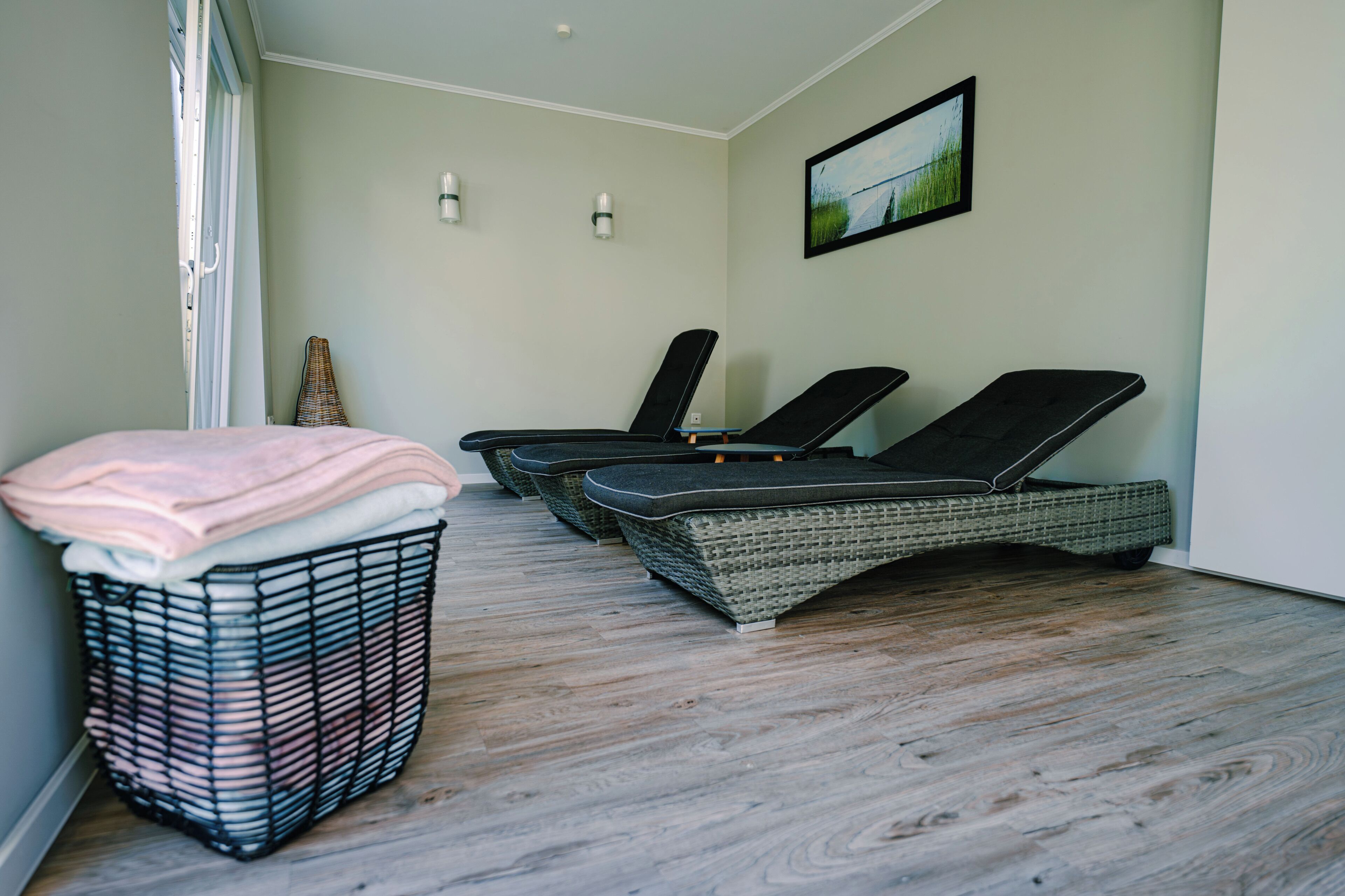 A room with a wicker lounge chair, a wicker basket with towels, and a framed picture on the wall.