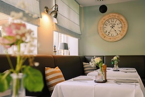 A restaurant interior with a dining table set for two, a wall clock, and a sofa with striped cushions. - Waastwinj (Utersum)